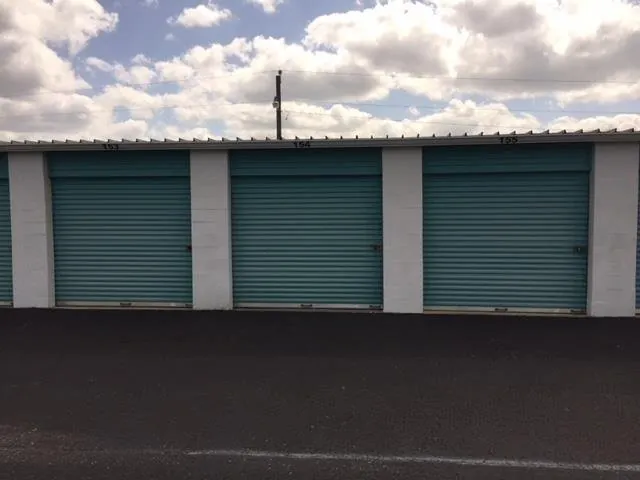Three adjacent drive-up storage units numbered 153, 154 and 155, their teal roll-up doors closed and latched at ground level with no step or kerb.