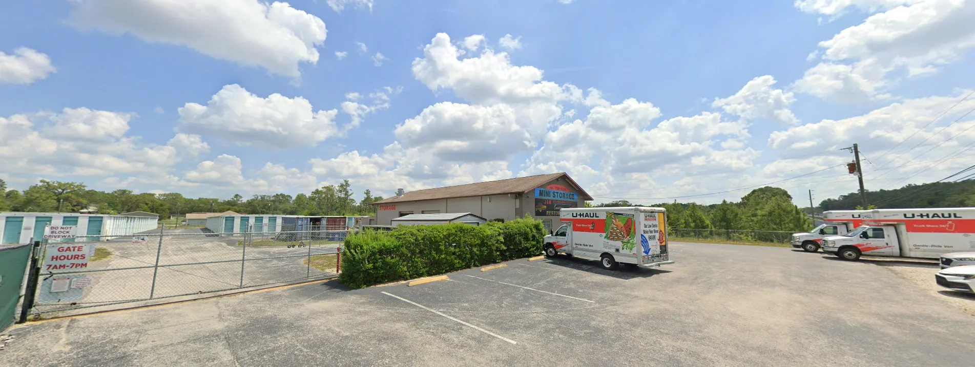 Wide view of the J&M Storage lot: a row of drive-up units with teal doors along the left, the office building at the centre, and two U-Haul rental trucks parked on the right. Signs at the entrance read 'Do not block driveway' and 'Gate hours 7am-7pm'.